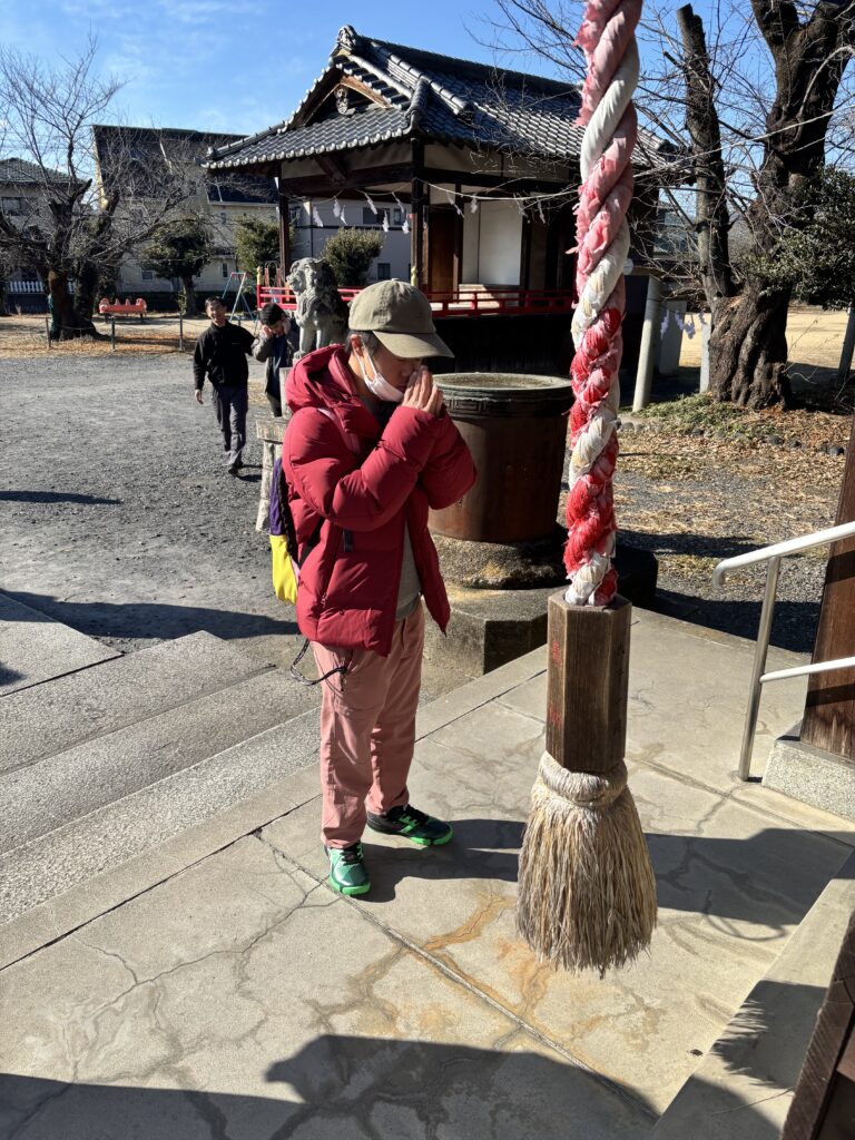 赤い上着を着た利用者さんが神社に手を合わせている様子を社殿側から撮影。空は青く、紅白の鈴緒が手前にある。