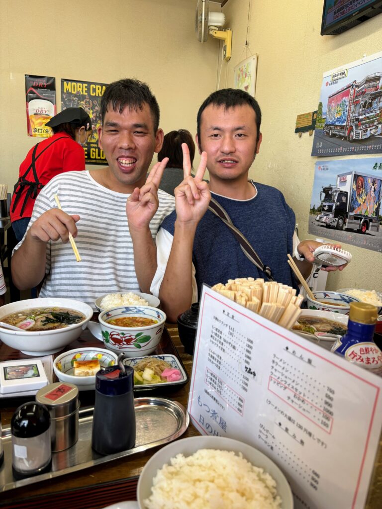もつ煮とラーメンを前にカメラ目線でピースする２０代の男性利用者さん二人